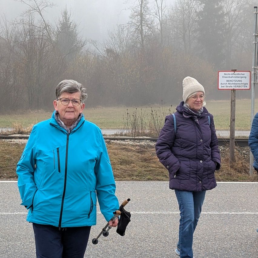 Zwei ältere Frauen gehen auf einer Straße mit nebligem Hintergrund, eine in einer blauen Jacke und die andere in einer lila Jacke, beide tragen Winterkleidung und Brillen.