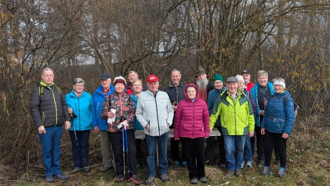 Eine Gruppe älterer Personen in Winterkleidung steht in einer Waldlichtung, einige halten Stöcke. Hinter ihnen steht eine Holzbank vor einem Hintergrund von laublosen Bäumen.