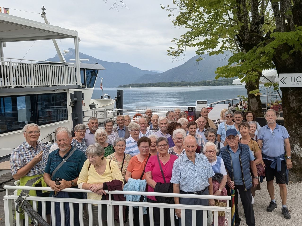 Eine Gruppe älterer Menschen steht auf einem Steg und posiert für ein Foto mit einem Boot und einem See im Hintergrund.