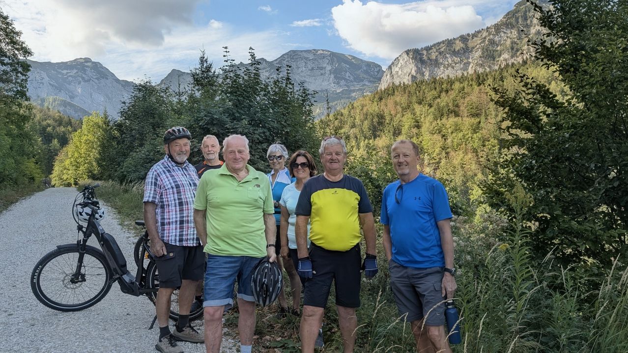 Eine Gruppe älterer Männer und Frauen steht lächelnd auf einem Pfad in den Bergen für ein Foto. Sie sind in Fahrradausrüstung gekleidet und einige tragen Sonnenbrillen. Hinter ihnen ist ein üppiger grüner Hang und Berge unter einem teilweise bewölkten Himmel sichtbar.
