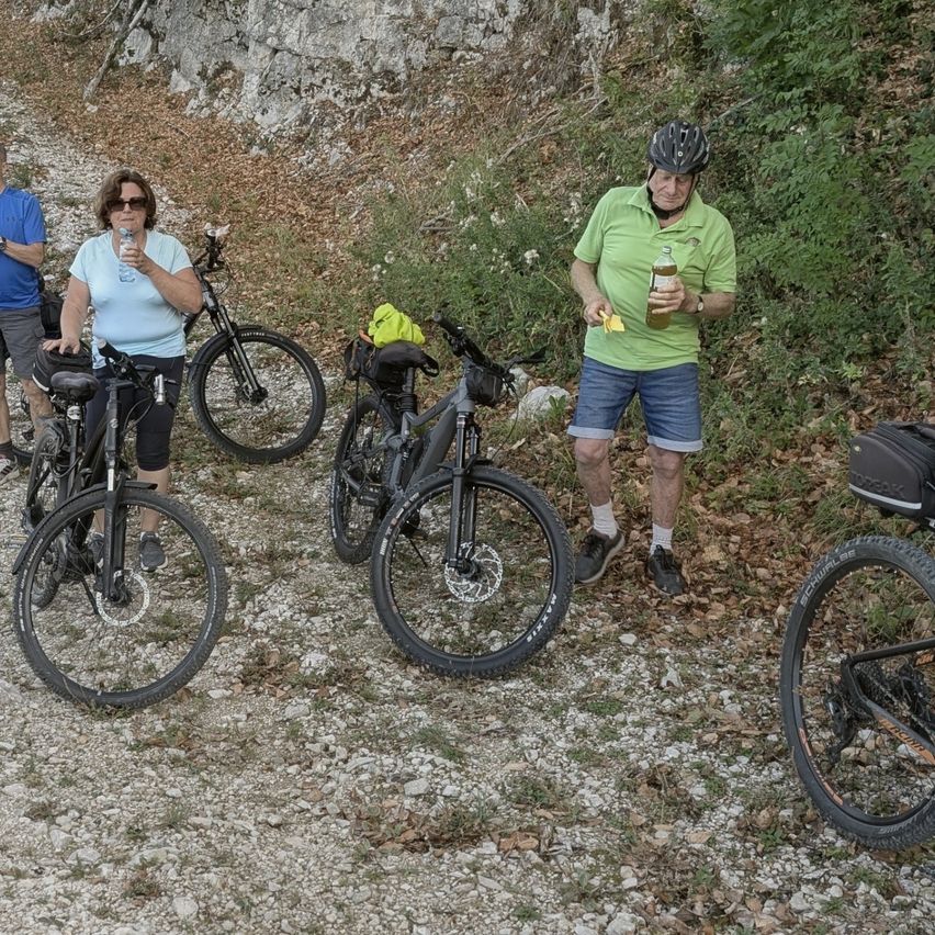 Eine Gruppe von Menschen steht auf einem felsigen Pfad mit ihren Fahrrädern. Eine Frau in einem blauen Shirt hält eine Flasche, während ein Mann in einem grünen Shirt eine Flasche und einen gelben Gegenstand hält.