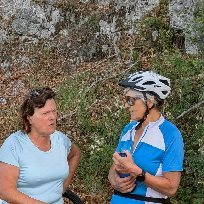 Zwei Frauen, eine mit Fahrradhelm, stehen in einer natürlichen Umgebung, eine hält eine blaue Flasche.