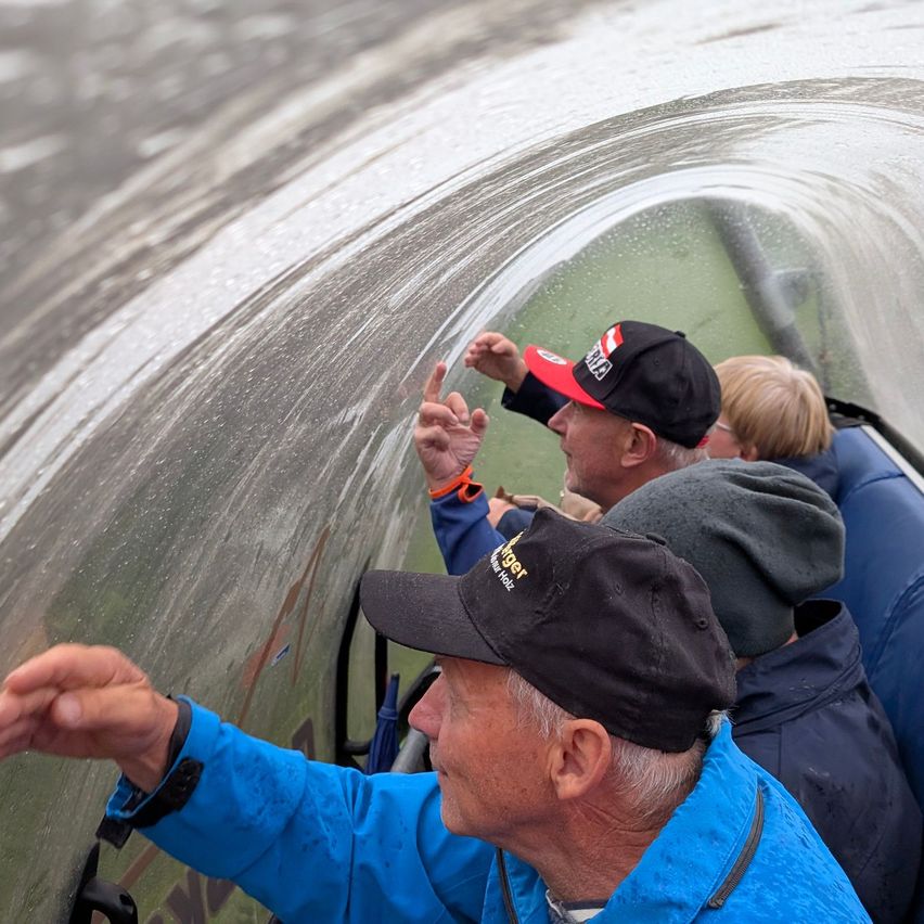 Eine Gruppe von Menschen in einem Boot, wobei ein Mann auf das Wasser zeigt. Sie tragen Hüte und Jacken.