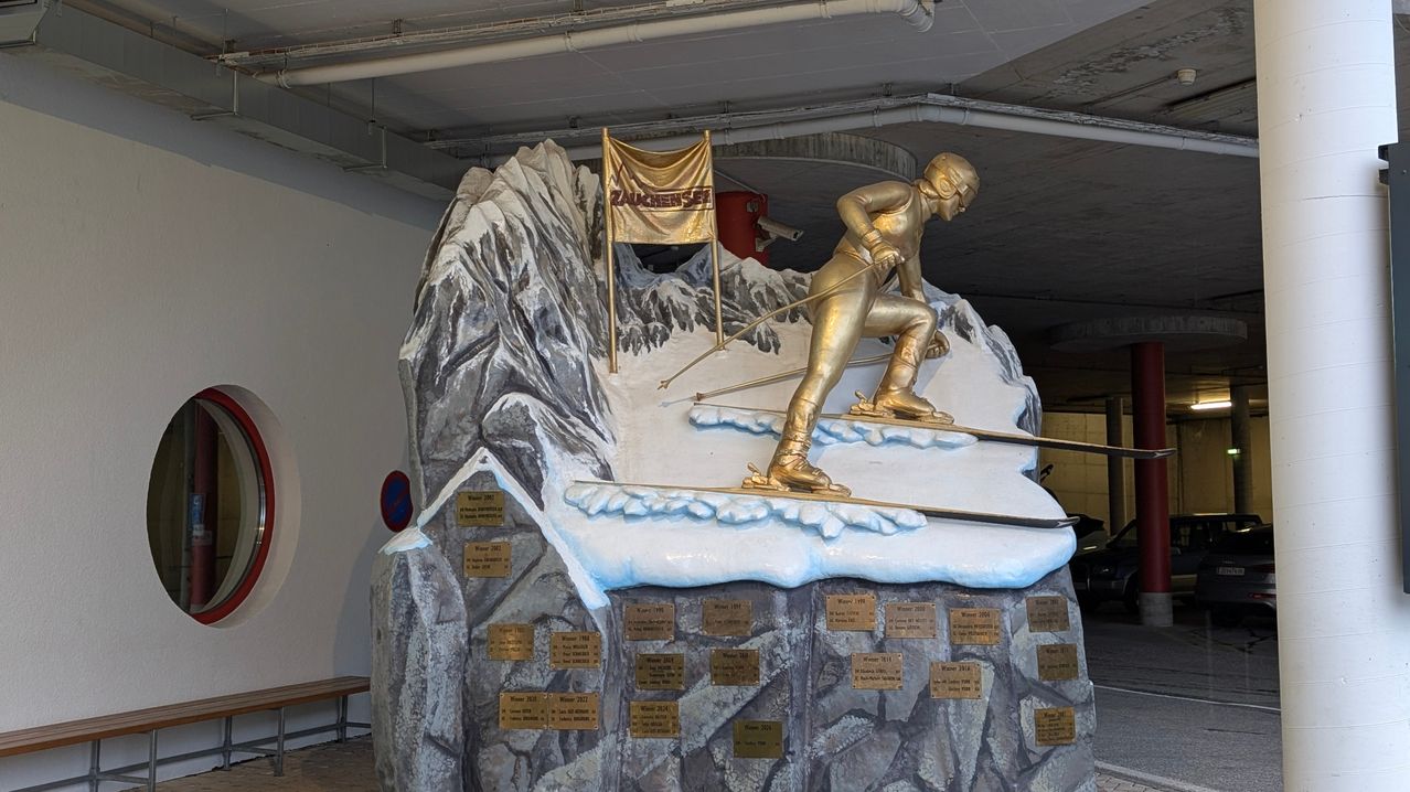 Eine goldene Statue eines Skifahrers auf einem verschneiten Berg mit Plaketten darunter. Der Skifahrer hält Skistöcke und trägt Skiausrüstung. Hinter dem Skifahrer ist eine Fahne mit dem Wort 'Zuckersee'.