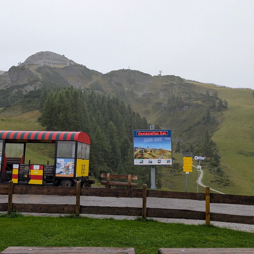Ein kleiner Zug auf einer Bahnstrecke mit einem Schild dahinter. Das Gebiet ist von einem Holzzaun und grünem Gras umgeben. Im Hintergrund befindet sich ein Berg mit Bäumen und einem nebligen Himmel.
