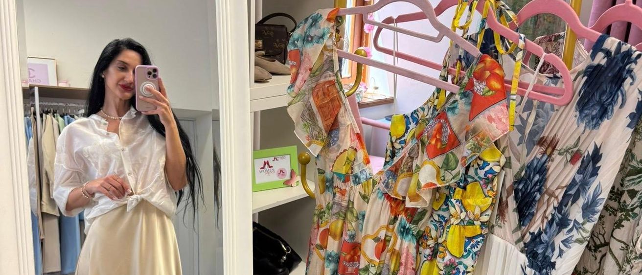 Eine Frau macht ein Selfie vor einem Spiegel, der einen Bekleidungsladen mit Blumenkleidern an Kleiderbügeln widerspiegelt. Die Regale hinter ihr halten Taschen und Bilderrahmen.