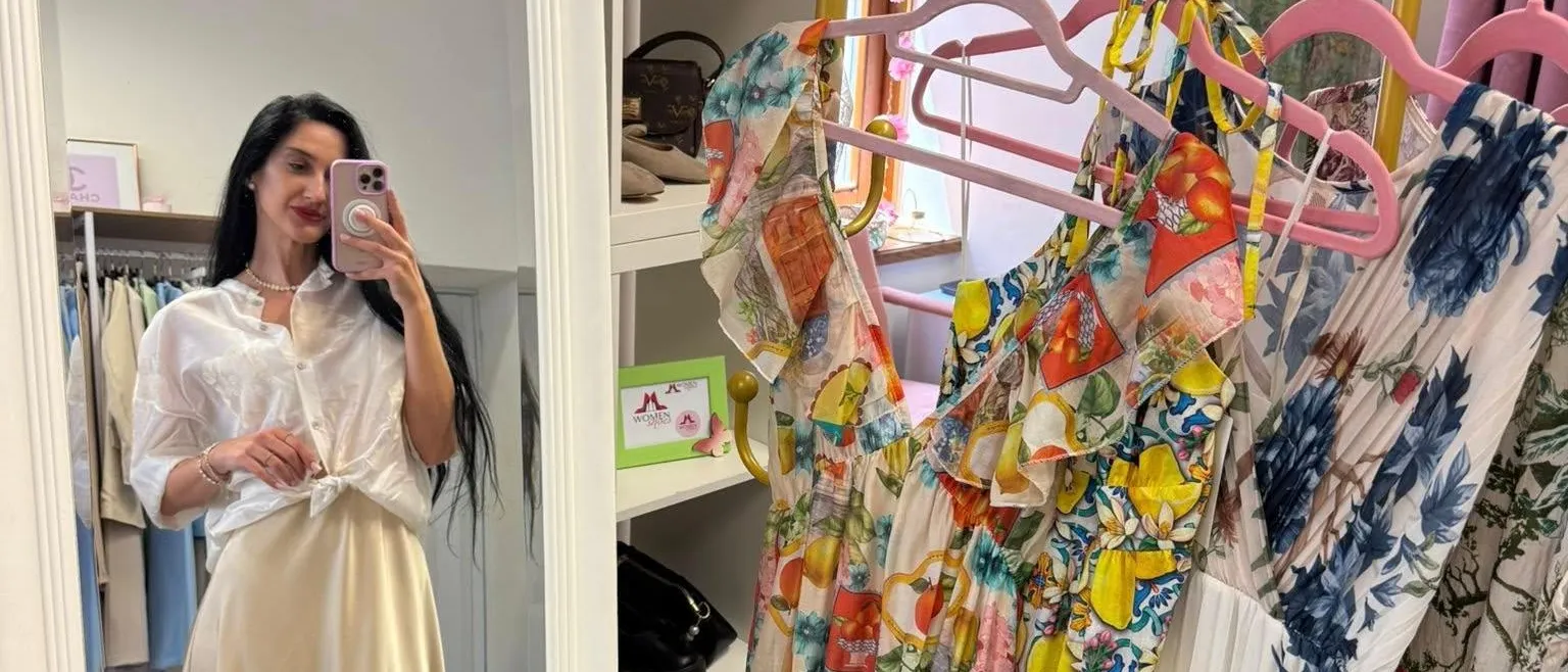 Eine Frau macht ein Selfie vor einem Spiegel, der einen Bekleidungsladen mit Blumenkleidern an Kleiderbügeln widerspiegelt. Die Regale hinter ihr halten Taschen und Bilderrahmen.