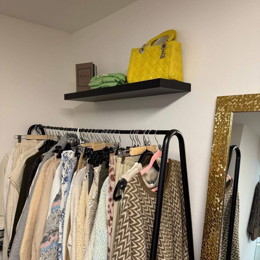 Ein Kleiderschrank mit verschiedenen Kleidungsstücken und einer gelben Gucci-Tasche auf einem Regal darüber. Ein Spiegel mit goldenen Akzenten steht rechts.