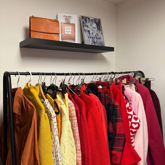 Ein Kleiderschrank mit verschiedenen Kleidungsstücken und Accessoires, darunter eine Chanel-Parfümflasche und ein Buch mit dem Titel 'Vintage', alles an einem schwarzen Metallständer aufgehängt.