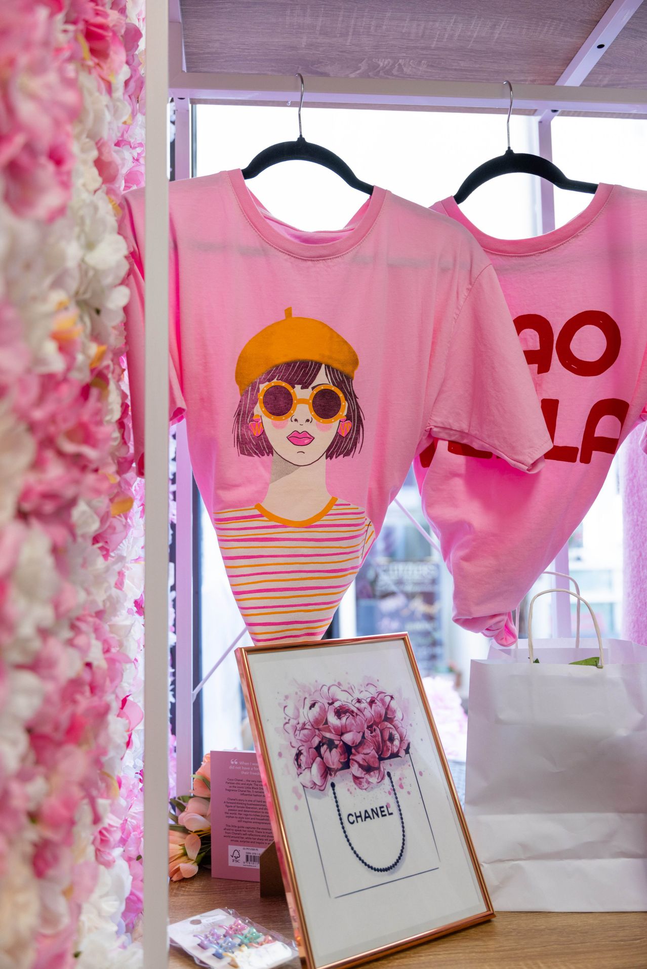 Zwei rosa T-Shirts hängen an Kleiderbügeln. Eines zeigt ein Porträt einer Frau mit Sonnenbrille, das andere steht 'DOLLA'. Ein Bilderrahmen und weiße Taschen sind in der Nähe.