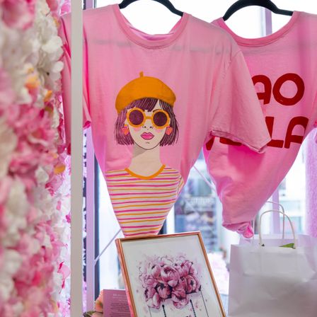 Zwei rosa T-Shirts hängen an Kleiderbügeln. Eines zeigt ein Porträt einer Frau mit Sonnenbrille, das andere steht 'DOLLA'. Ein Bilderrahmen und weiße Taschen sind in der Nähe.