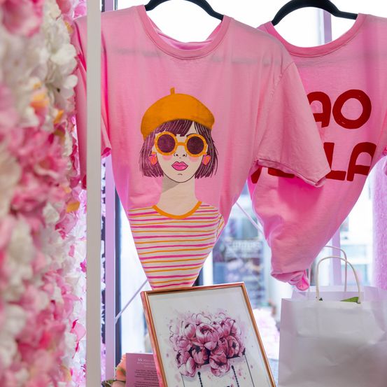 Zwei rosa T-Shirts hängen an Kleiderbügeln. Eines zeigt ein Porträt einer Frau mit Sonnenbrille, das andere steht 'DOLLA'. Ein Bilderrahmen und weiße Taschen sind in der Nähe.