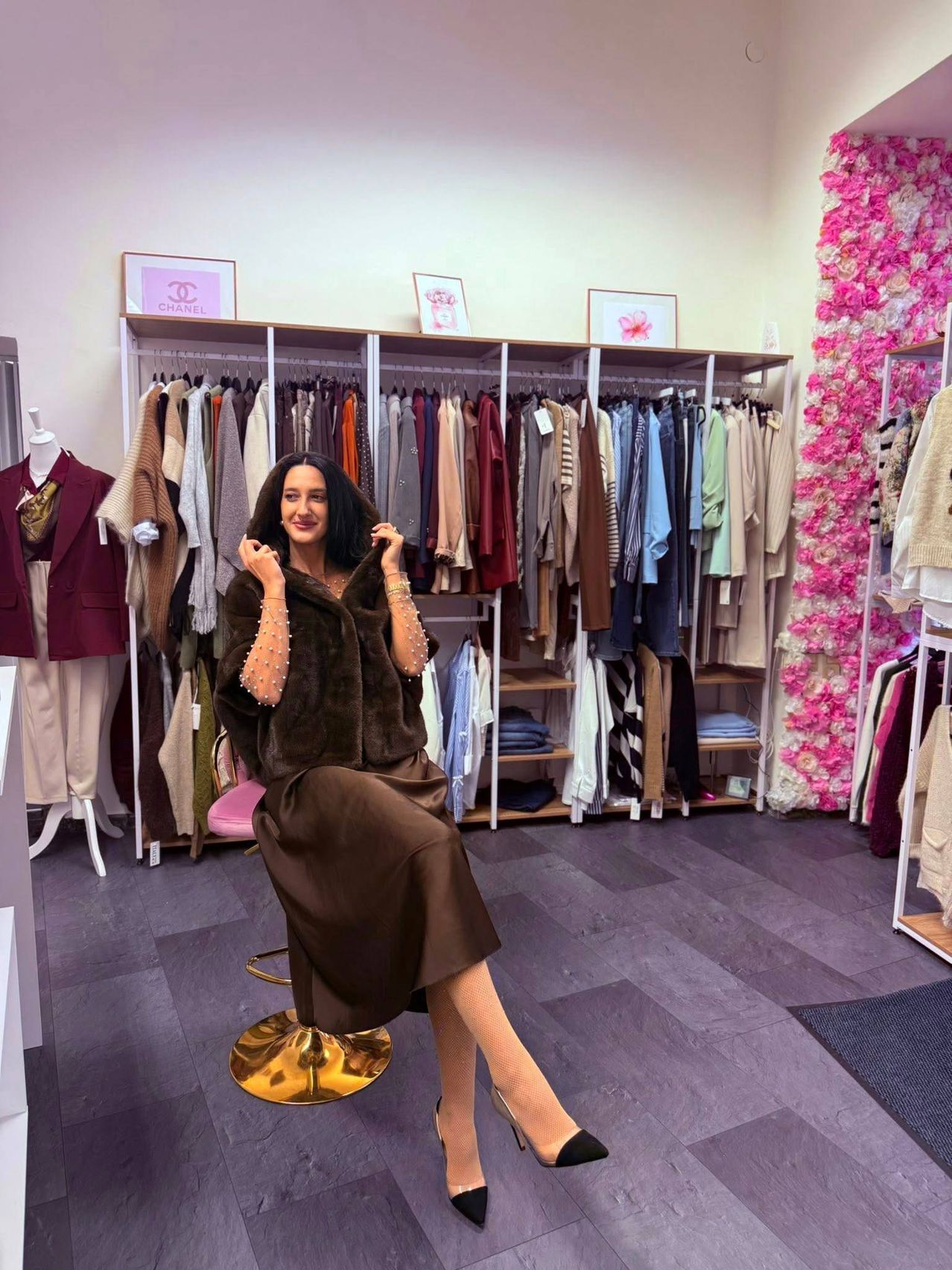 Eine Frau sitzt auf einem Stuhl in einem Bekleidungsgeschäft und lächelt. Hinter ihr hängt ein Kleiderständer, und Chanel-Schilder sind an der Wand zu sehen.