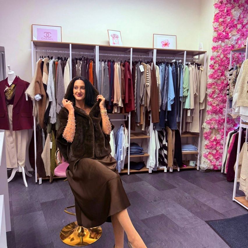 Eine Frau sitzt auf einem Stuhl in einem Bekleidungsgeschäft und lächelt. Hinter ihr hängt ein Kleiderständer, und Chanel-Schilder sind an der Wand zu sehen.