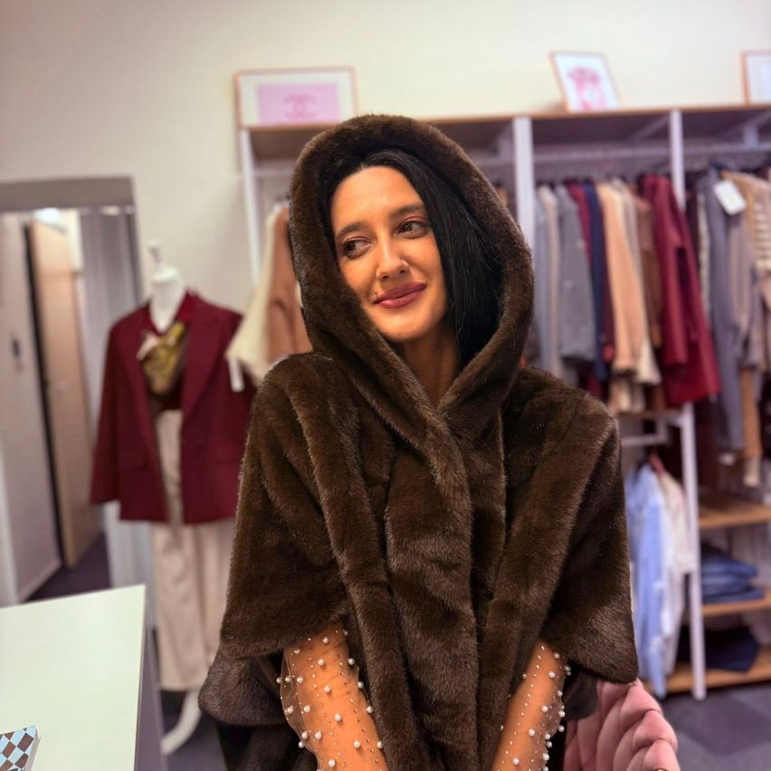 Eine Frau in einem braunen Pelzmantel mit Kapuze lächelt in einem Bekleidungsgeschäft, umgeben von Kleiderständern.