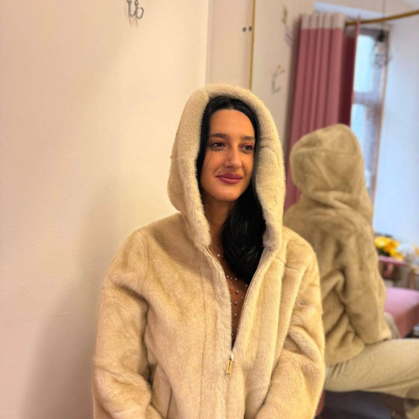 Eine Frau in einem cremefarbenen Kapuzenpelzmantel lächelt und steht an einer weißen Wand, mit einer weiteren Person in einem ähnlichen Mantel im Hintergrund.