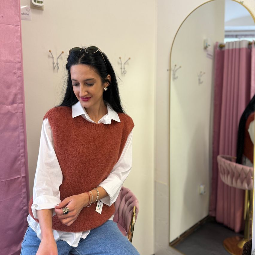 Eine Frau in einem braunen Pullover, einem weißen Hemd und blauen Jeans sitzt auf einem Stuhl und posiert für ein Foto. Hinter ihr spiegelt ein Spiegel einen rosa Vorhang und einen Samtstuhl wider.