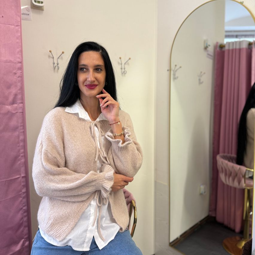 Eine Frau in einem weißen Hemd und einem beigen Cardigan posiert vor einem Spiegel mit rosa Vorhängen.