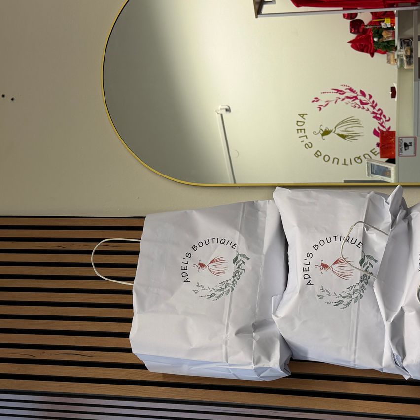 Drei weiße Adel's Boutique Papiertüten liegen auf einer Holzbank vor einem Spiegel mit dem Adel's Boutique Logo an der Wand.