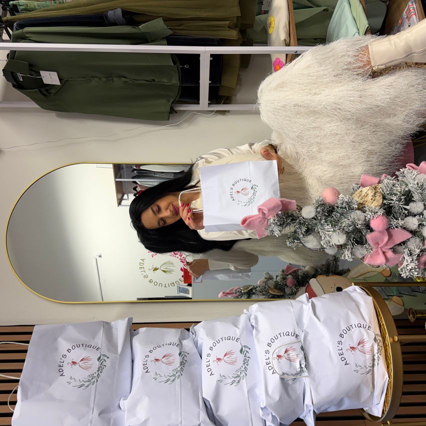 Eine Frau hält einen Papierbeutel vor einem Spiegel mit einem Weihnachtsbaum neben ihr, umgeben von anderen Papiertüten in einem Boutique.