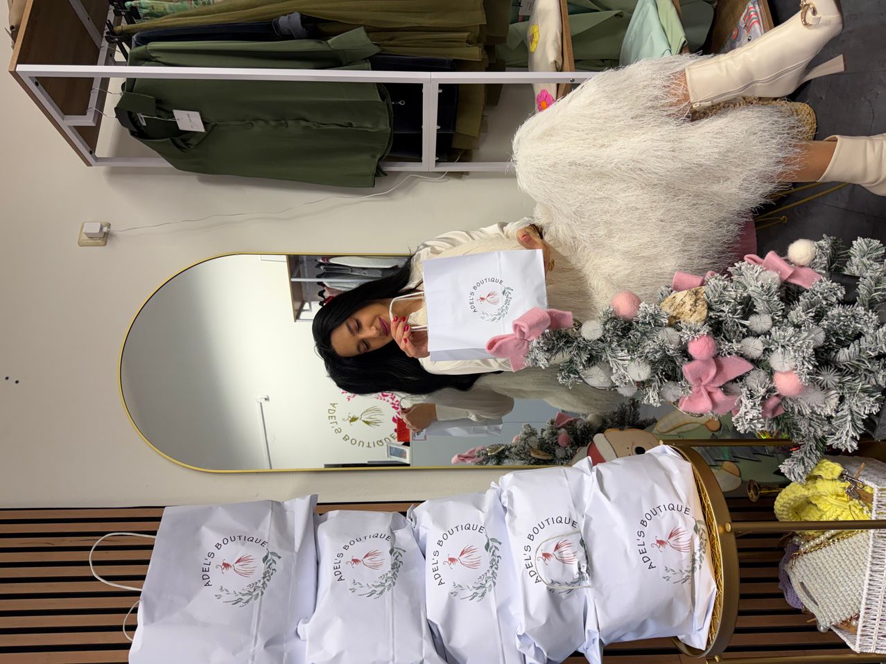 Eine Frau hält einen Papierbeutel vor einem Spiegel mit einem Weihnachtsbaum neben ihr, umgeben von anderen Papiertüten in einem Boutique.