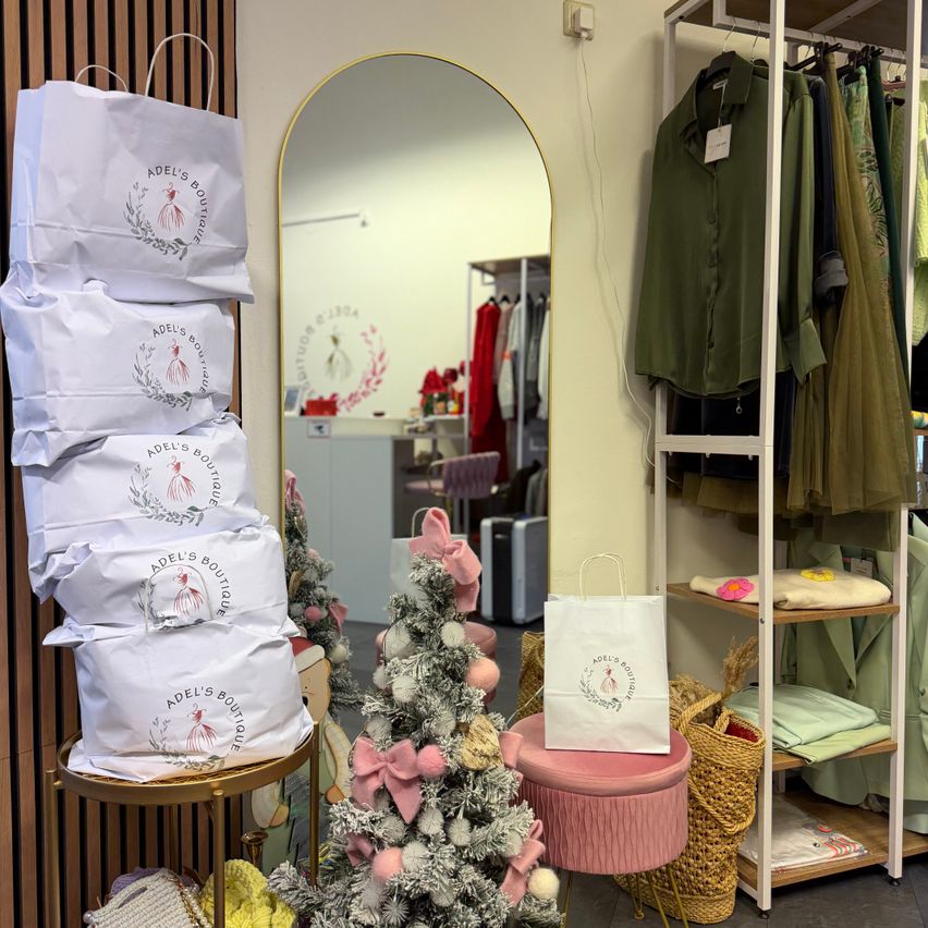 Ein Boutique-Interieur mit einem Weihnachtsbaum, einem großen Spiegel und Kleidung an Kleiderbügeln. Mehrere weiße Taschen mit der Aufschrift Adel's Boutique sind auf einem Tisch gestapelt. Ein rosa Hocker hält eine weiße Tasche.