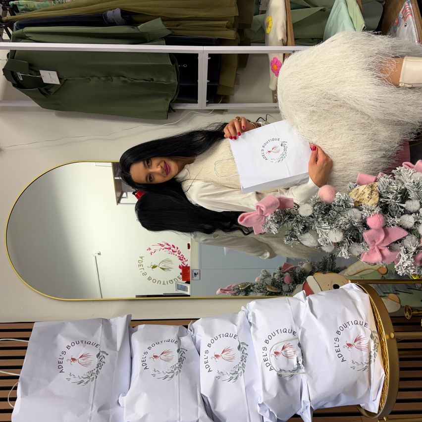 Eine Frau in einem Pelzkleid posiert mit einer Tasche vor einem Spiegel. Sie hält ein Papier mit dem Logo Adel's Boutique. Es gibt festliche Dekorationen, einschließlich eines Weihnachtsbaums, und mehrere Taschen mit demselben Logo.