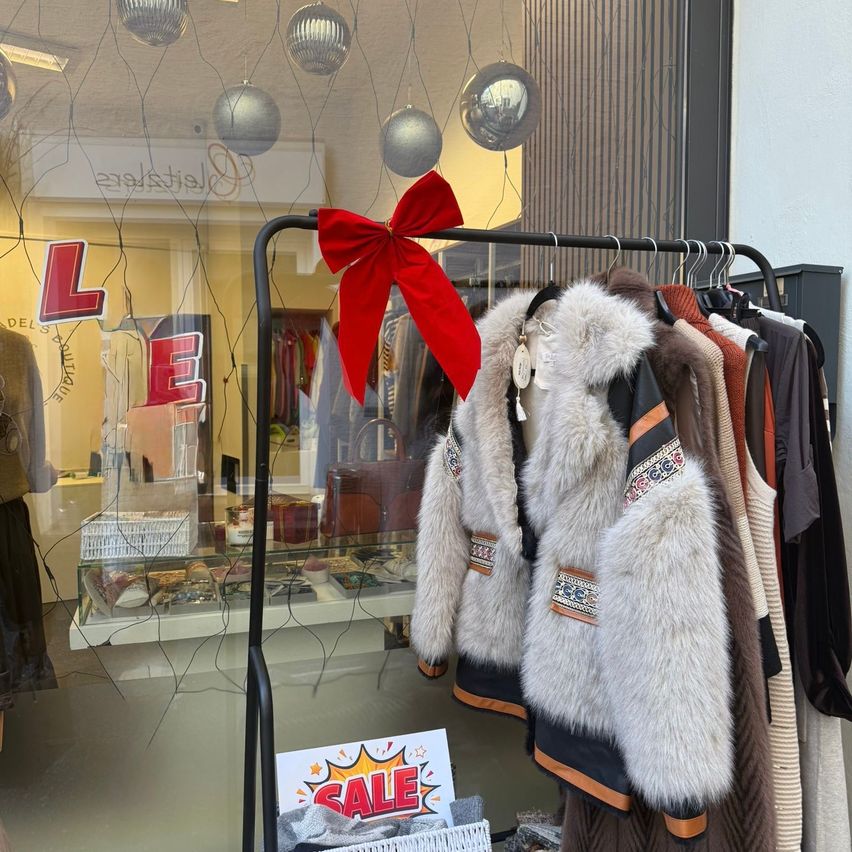 Ein Bekleidungsgeschäft zeigt Wintermäntel an einem Kleiderständer mit einer roten Schleife. Das Schaufenster des Ladens zeigt ein Verkaufsschild und Artikel im Inneren.