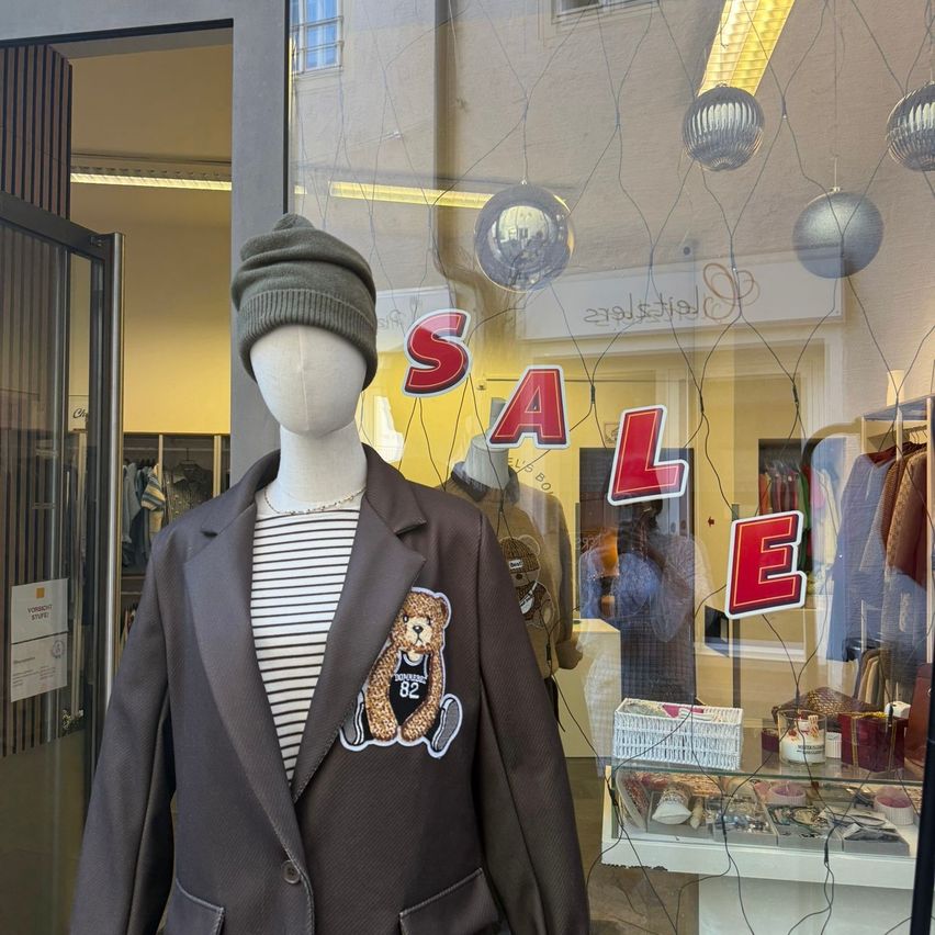 Eine Schaufensterpuppe trägt ein gestreiftes Shirt und einen braunen Blazer mit einem Bären-Patch. Das Wort 'SALE' ist im Schaufenster zu sehen.