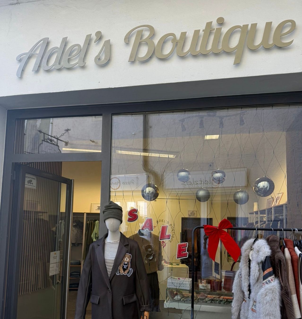 Ein Boutique-Schaufenster mit dem Schild Adel's Boutique. Ein Mannequin in einem braunen Mantel ist vor einem Glasfenster ausgestellt. Ein roter Schleifenband schmückt einen Kleiderständer mit verschiedenen Mänteln.