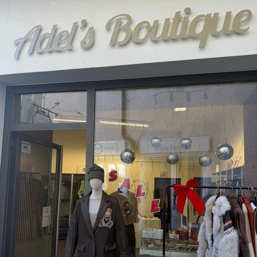 Ein Boutique-Schaufenster mit dem Schild Adel's Boutique. Ein Mannequin in einem braunen Mantel ist vor einem Glasfenster ausgestellt. Ein roter Schleifenband schmückt einen Kleiderständer mit verschiedenen Mänteln.