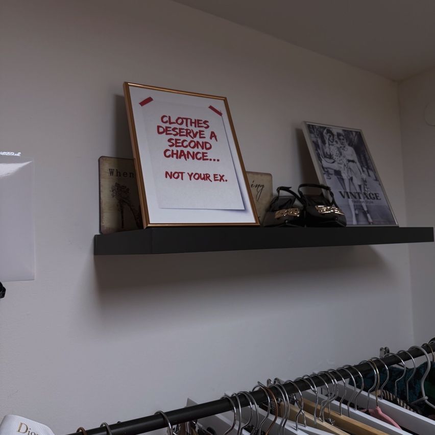 An einer weißen Wand ist ein schwarzes Regal mit einem gerahmten Zitat und einem Bild, einem kleinen Schild und einem Paar Vintage-Schuhe zu sehen. Unter dem Regal befindet sich ein Kleiderständer mit zahlreichen Kleiderbügeln.