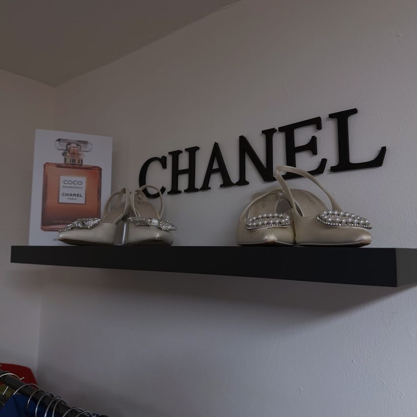 Eine weiße Wand mit einem schwarzen Regal hält zwei Chanel-Schuhe und eine Coco Chanel-Parfümflasche.