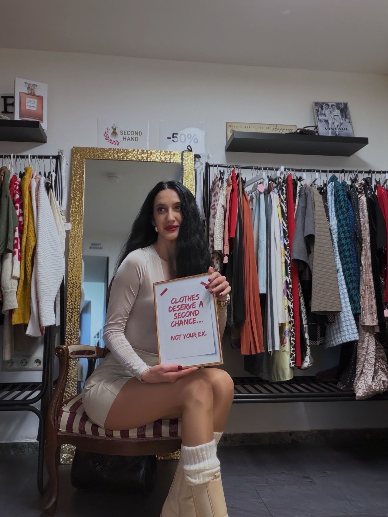 Eine Frau in einem weißen Kleid sitzt auf einem Stuhl und hält ein Schild mit der Aufschrift 'Kleider verdienen eine zweite Chance... nicht deine Ex.' Sie steht vor einem Bekleidungsgeschäft mit einem Spiegel und einem Kleiderständer.
