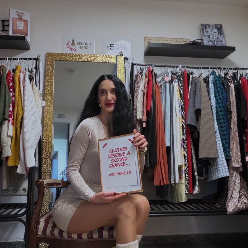 Eine Frau in einem weißen Kleid sitzt auf einem Stuhl und hält ein Schild mit der Aufschrift 'Kleider verdienen eine zweite Chance... nicht deine Ex.' Sie steht vor einem Bekleidungsgeschäft mit einem Spiegel und einem Kleiderständer.