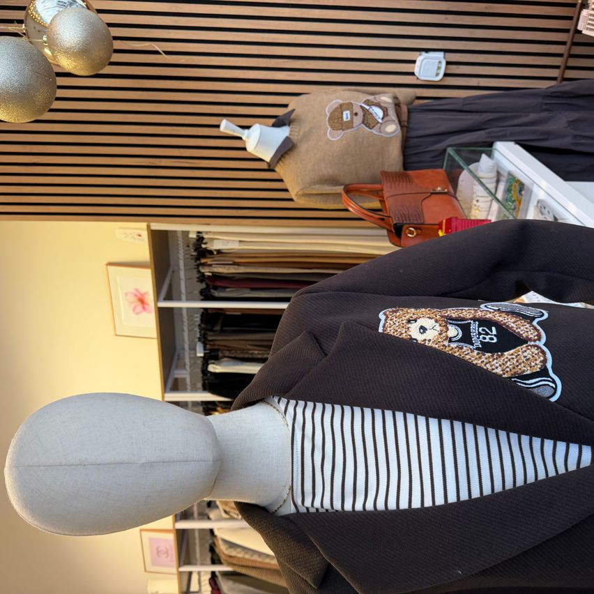 Ein Mannequin zeigt einen braunen Blazer über einem gestreiften Shirt, mit einem braunen Teddybär-Kissen und einer Lederhandtasche dahinter. Eine dekorative silberne Kugel hängt von der Decke ab.
