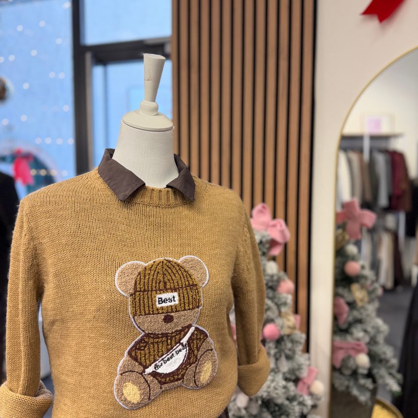 Ein beiger Pullover mit braunem Kragen und einer Teddybär-Stickerei wird auf einem Mannequin in einem Geschäft ausgestellt. Der Hintergrund zeigt einen Weihnachtsbaum und ein Fenster.