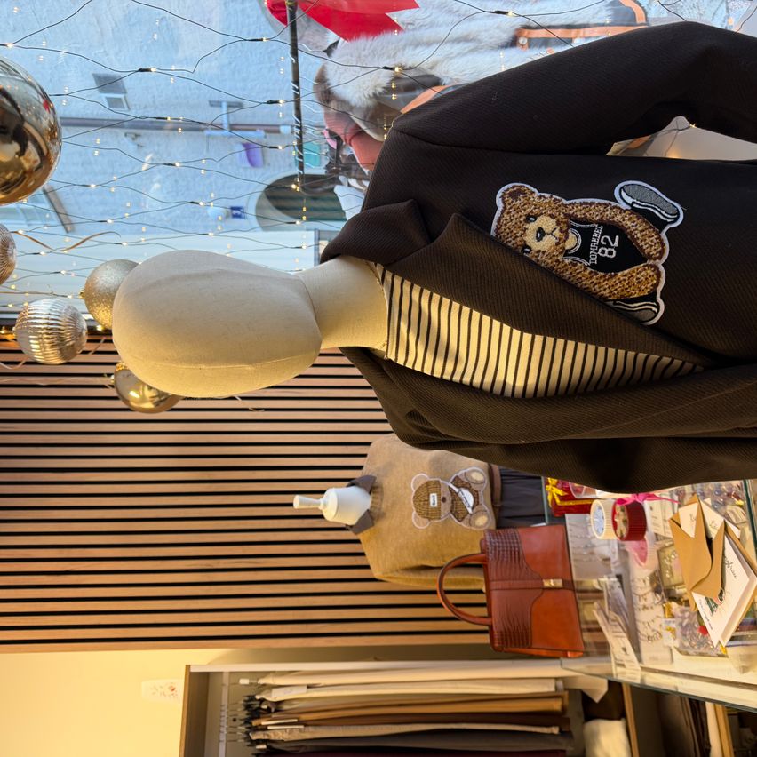 Ein Mannequin zeigt einen schwarzen Blazer mit einem Bären-Grafik und gestreiftes Shirt darunter, vor einem Hintergrund von dekorativen Lichtern und Taschen.