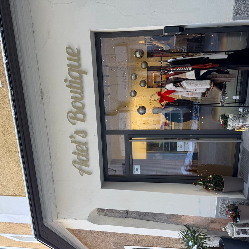 Ein Blick von oben auf Adel's Boutique, mit einem Glasfenster, durch das Kleidungsstücke zu sehen sind. Der Name des Geschäfts ist über dem Fenster auf der Wand angebracht.