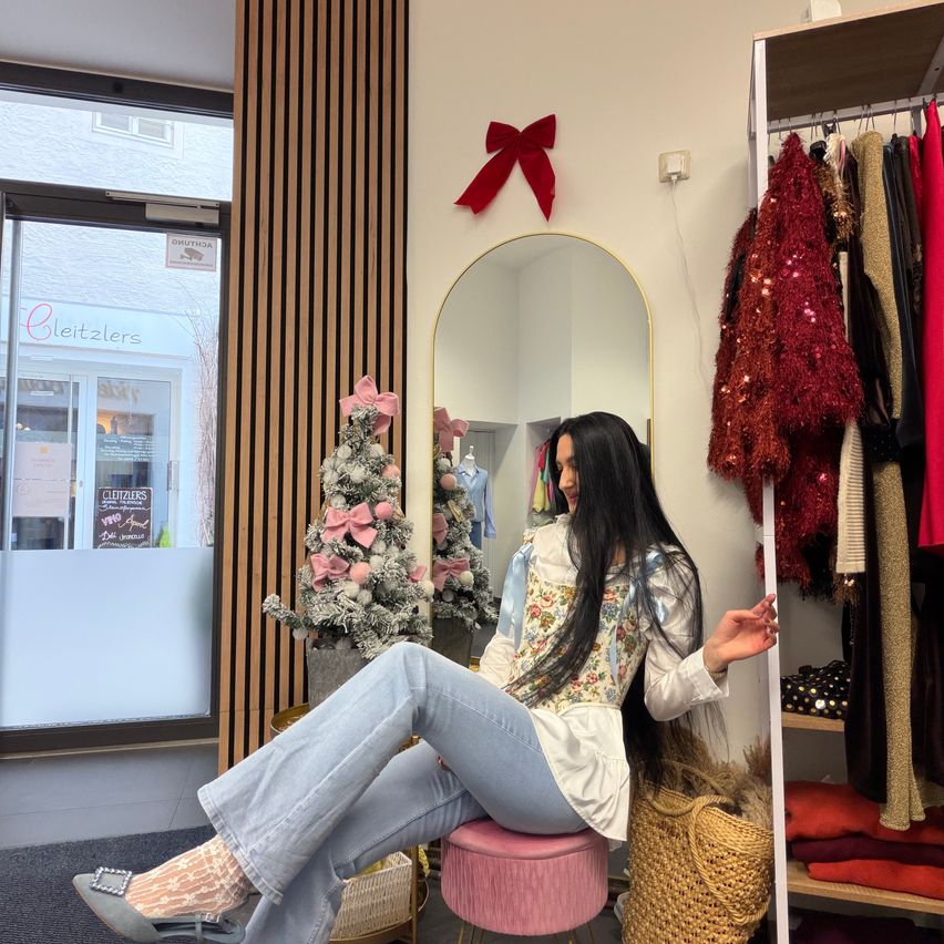 Eine Frau sitzt auf einem pinken Hocker in einem Bekleidungsgeschäft und posiert vor einem Spiegel. Hinter ihr ist ein Weihnachtsbaum mit rosa Schleifen zu sehen. Das Geschäft zeigt verschiedene Kleidungsstücke an Kleiderbügeln. Das Schild des Geschäfts lautet 'Cleitzlers'.