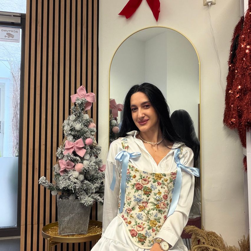 Eine Frau steht vor einem Spiegel und lächelt. Sie trägt ein Blumenkleid mit blauen Bändern. Hinter ihr steht ein Weihnachtsbaum mit rosa Schleifen auf einem Tisch. Eine rote Stoffbahn hängt an der Wand.