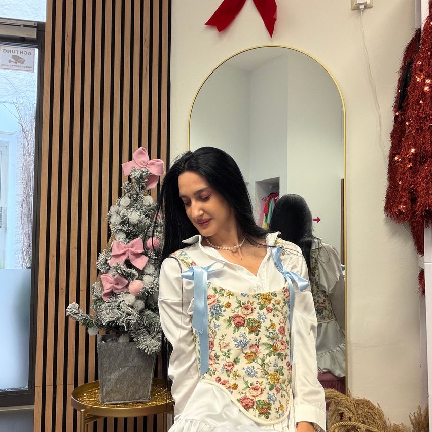 Eine Frau steht vor einem Spiegel und lächelt. Sie trägt ein Blumenkleid mit blauen Bändern und eine Halskette. Hinter ihr steht ein Weihnachtsbaum mit rosa Schleifen und ein Schild an der Wand.