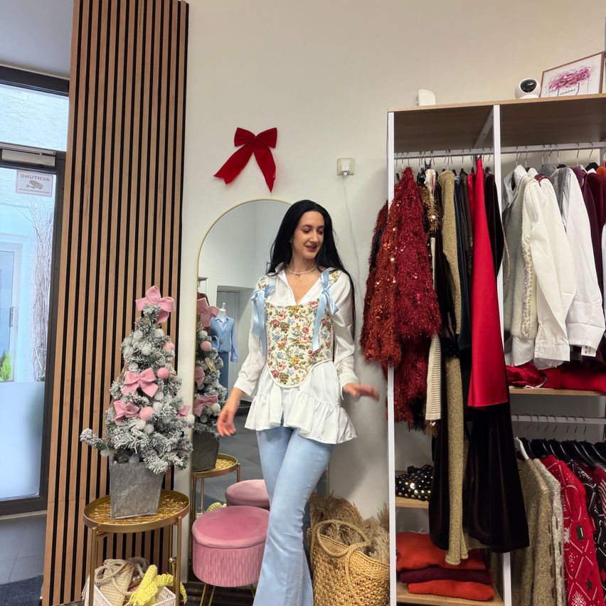 Eine Frau in einer weißen Bluse und blauen Hosen steht in einem Bekleidungsgeschäft und lächelt, mit einem Weihnachtsbaum und einem Regal mit verschiedenen Kleidungsstücken hinter ihr.