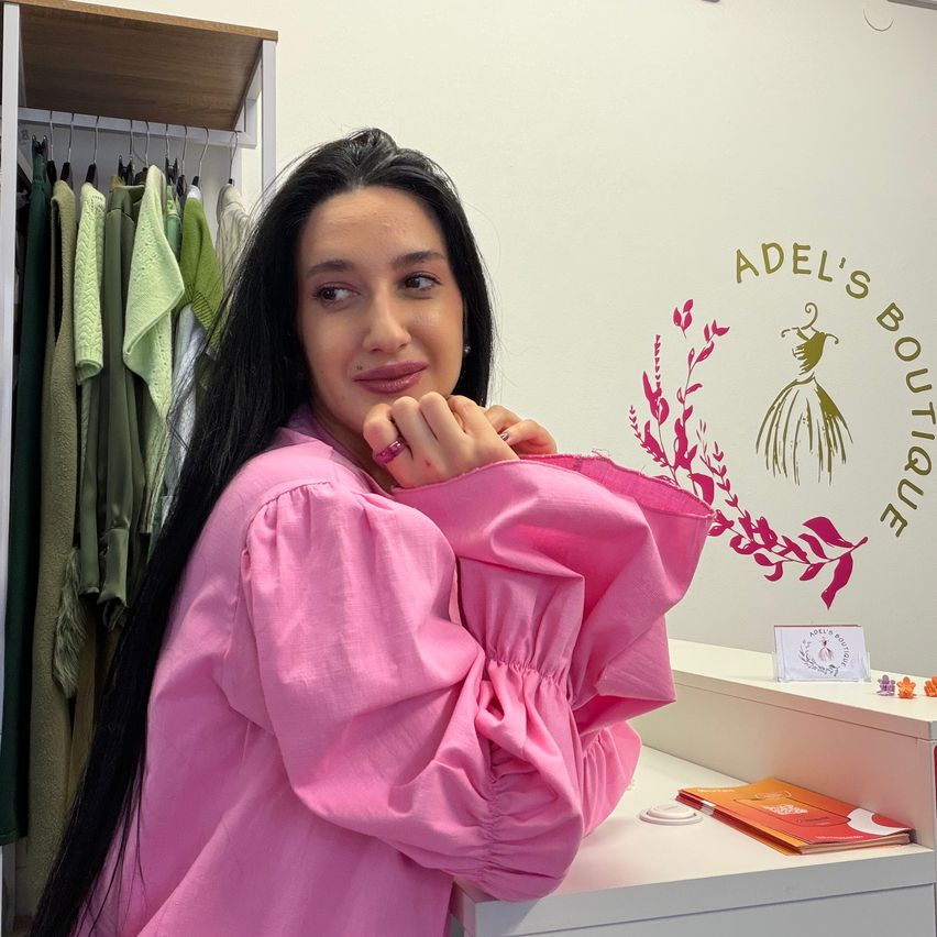 Eine Frau mit langen schwarzen Haaren steht vor einem weißen Regal mit Kleidung. Sie trägt ein rosa Langarm-Hemd mit gepufften Ärmeln und einen Ring am Finger. Hinter ihr befindet sich eine Wand mit einem Logo und einigen kleinen Figuren.