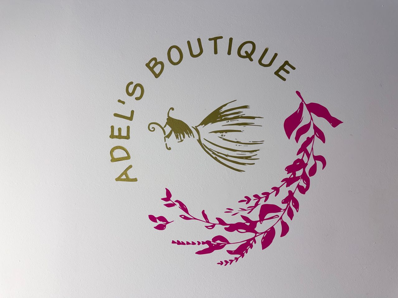 Das Bild zeigt das Logo von Adel's Boutique mit goldenem Text und einem stilisierten Blatt sowie einem Schmetterlingsdesign in Pink.