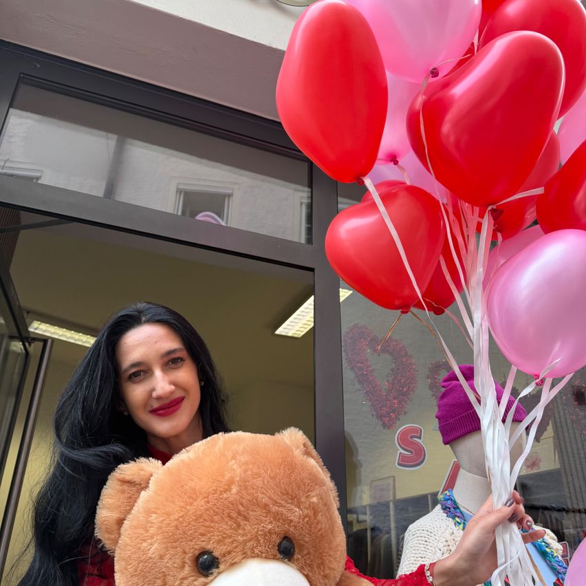 Eine Frau hält einen Strauß herzförmiger Ballons und einen braunen Teddybären vor einem Geschäft. Das Schild des Geschäfts ist im Hintergrund sichtbar.