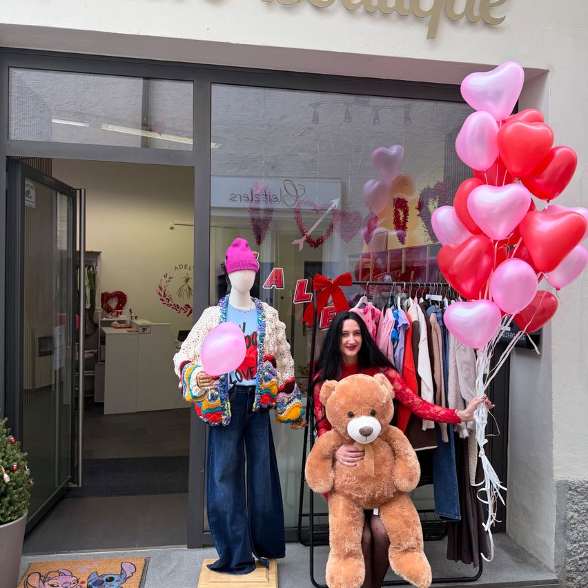 Eine Frau steht vor einem Bekleidungsgeschäft, hält einen Teddybären und einen Strauß herzförmiger Luftballons und ein Mannequin mit einer rosa Mütze in der Nähe.