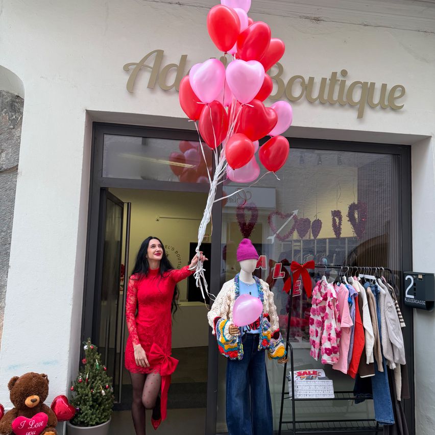 Eine Frau in einem roten Kleid hält einen Strauß roter und rosa Herzballons vor einem Boutiqueladen mit einem Dummy, das eine pinke Mütze trägt. Der Laden zeigt Kleidung, und Dekorationen umfassen einen Weihnachtsbaum und einen Bären.