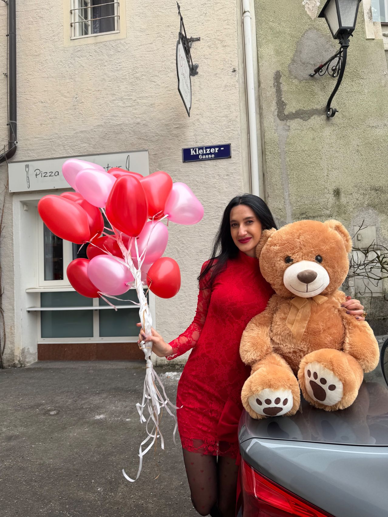 Eine Frau in einem roten Kleid hält einen Strauß roter und rosa Herzballons und einen großen braunen Teddybären vor einem Gebäude mit einem Schild, das 'Pizza' steht.