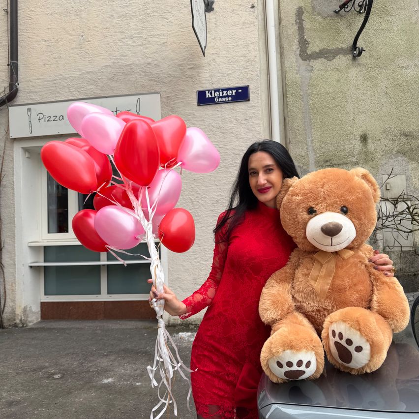 Eine Frau in einem roten Kleid hält einen Strauß roter und rosa Herzballons und einen großen braunen Teddybären vor einem Gebäude mit einem Schild, das 'Pizza' steht.